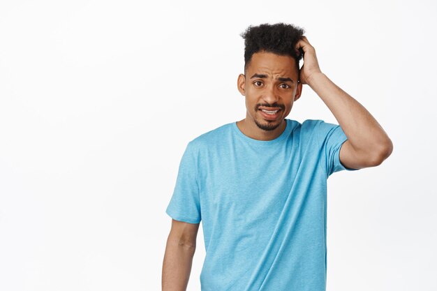 Awkward worried african american man scratch head guilty, furrow eyebrows and looking confused, puzzled to answer question, standing against white background