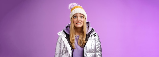 Free photo awkward worried cute timid blond girl in silver jacket hoodie winter hat clench teeth popping eyes