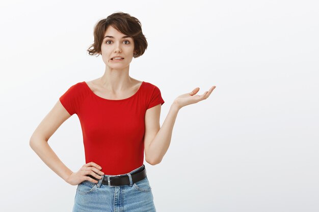 Awkward young woman raising hand confused and looking clueless