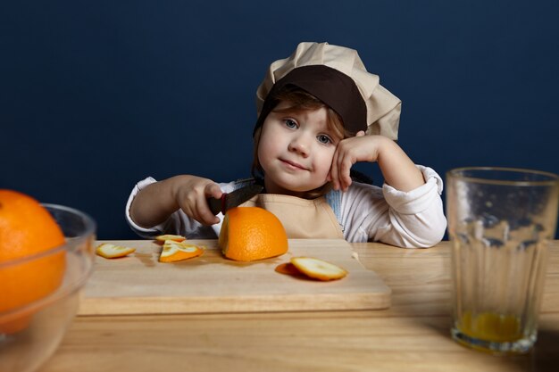 Baby chef in stylish apron and hat standing at table with wooden cutting board, using sharp knife while slicing fresh oranges for salad. Picture of adorable little girl helping mother in the kitchen