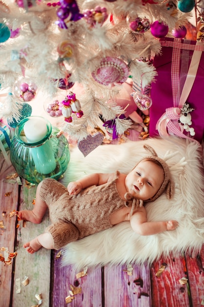 Baby in deer suit lies on soft white pillow under white Christmas tree with pink toys 