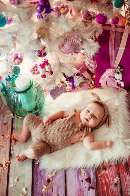 Baby in deer suit lies on soft white pillow under white Christmas tree with pink toys 