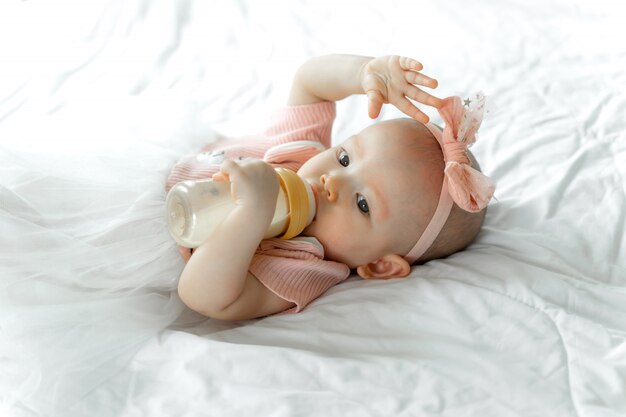 Baby drinks milk from a bottle on a white bed