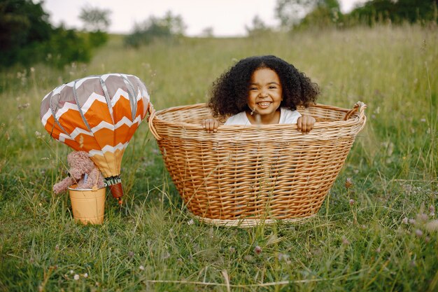 Девочка в плетеной корзине с воздушными шарами в солнечном свете в летнее время. Счастливый ребенок в поле на природе на траве