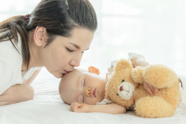 Baby sleeping with a teddy bear and mother kissing her