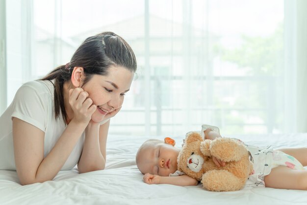 Baby sleeping with a teddy bear and mother looking at them