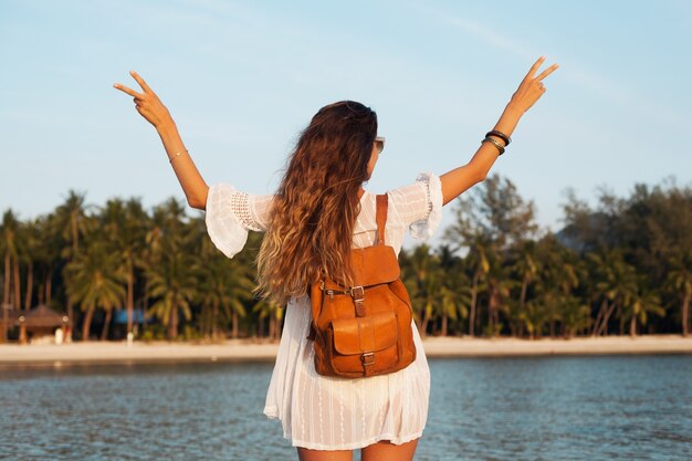 Back of beautiful woman in white dress walking carefree on tropical beach with leather backpack.