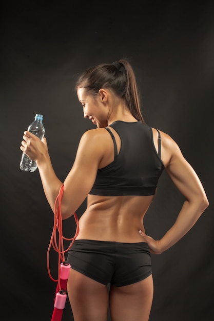 The back of muscular young woman athlete  with a skipping rope drinking water on black background.