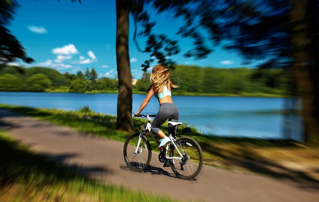 空気中の高い髪を飛んで湖の近くの緑の夏の公園で自転車に乗ってセクシーなホットスポーツブロンド女性少女モデルの裏