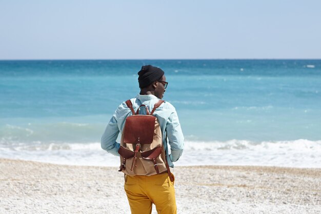 Back outdoor view of young African male tourist with knapsack wearing trendy clothing spending sunny morning at seaside, feeling happy and excited about seeing ocean for the first time in his life