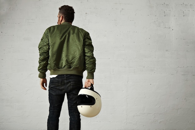 Free photo back portrait of a hip bearded pilot in green bomber jacket, skinny jeans and with a white blank helmet in his hand with white walls