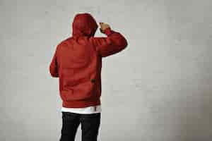 Free photo back portrait of a man in black jeans, white t-shirt and red parka putting on his hood on white wall