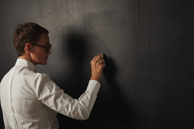 Back portrait of a serious female teacher in a white shirt starting to write onn empty blackboard
