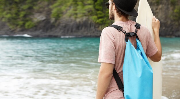 Foto gratuita colpo posteriore dell'uomo caucasico con la borsa blu che tiene tavola da surf, guardando i suoi amici navigare, cavalcando onde giganti in una ventosa giornata estiva