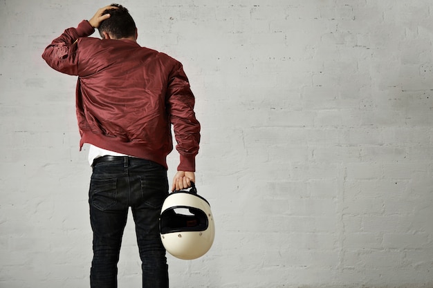 Бесплатное фото Задний снимок молодого мотоциклиста в джинсах, военной куртке-бомбардировщике и держащего белый шлем, касающегося его волос, изолированных на белом