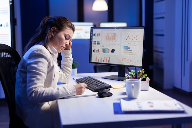 Free photo back shot of overwhelmed woman working at night in front of computer, writing notes on notebook annual reports, checking financial deadline