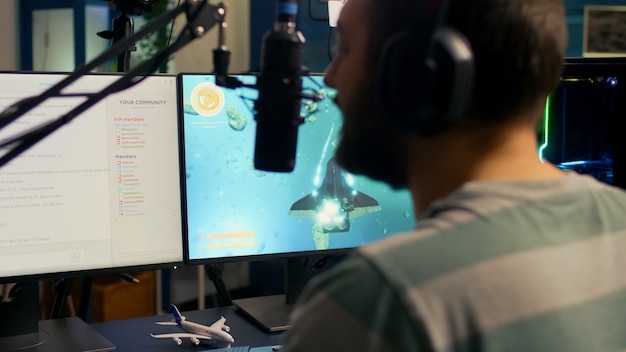 Back shot of streamer man playing on powerful computer shooter video game for tournament, talking with multiple players into headphones