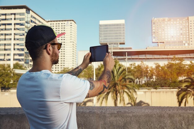 그의 태블릿에 도시 건물과 야자수의 사진을 찍는 평범한 흰색 티셔츠와 야구 모자에있는 젊은 남자의 백 샷.