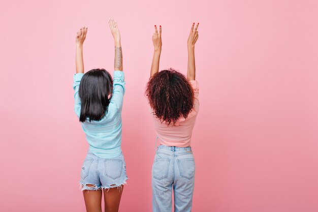 back of stretching brunette girls in jeans. Glamorous dark-haired ladies in vintage attire posing with hands up.