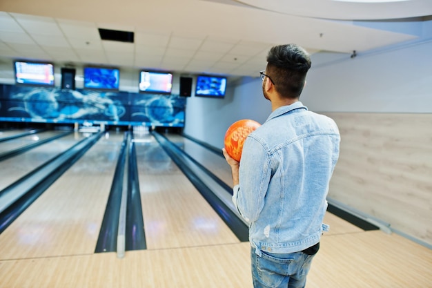 Retro dell'uomo asiatico alla moda in giacca di jeans e occhiali in piedi alla pista da bowling con la palla a portata di mano