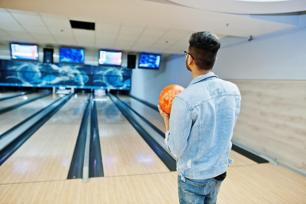 Back of stylish asian man in jeans jacket and glasses standing at bowling alley with ball at hand