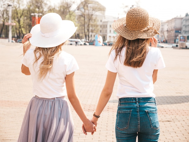 Назад двух молодых красивых белокурых улыбающихся хипстерских девочек в модной летней белой одежде футболки и шляпе. , Пара, держась за руки
