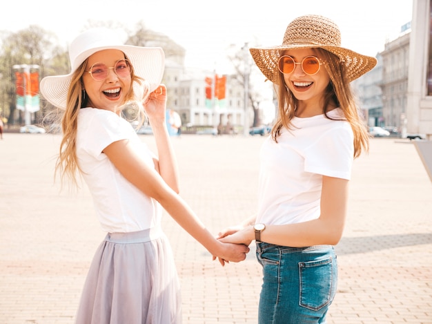 Back of two young beautiful blond smiling hipster girls in trendy summer white t-shirt clothes and hat.  . Couple holding each other hands