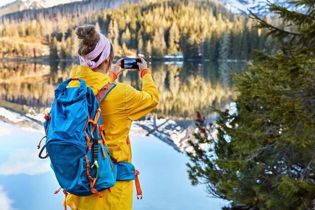 彼女のスマートフォンデバイス上の山と湖の風景のアクティブな女性の観光客の写真の背面図