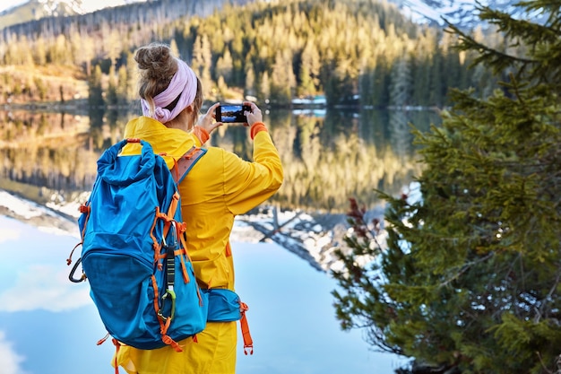 Вид сзади активной туристки фотографирует озерный пейзаж с горами на своем смартфоне