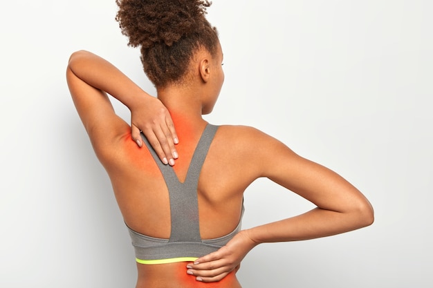 Back view of afro woman touches back and neck, shows inflammated zones, poses against white wall Free Photo