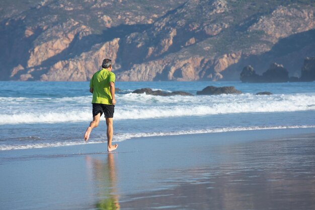 Back view of aged man training barefoot on seashore. Sporty man in T-shirt and shorts jogging in sea water strengthening health getting pleasure. Sport and healthy lifestyle of elderly people concept