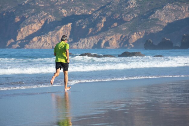Вид сзади на пожилого мужчину, тренирующегося босиком на берегу моря. Спортивный мужчина в футболке и шортах бегает в морской воде, укрепляя здоровье и получая удовольствие. Концепция спорта и здорового образа жизни пожилых людей