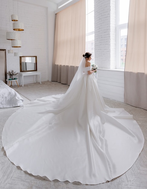 Вид сзади привлекательной невесты, одетой в роскошное свадебное платье, которая стоит в комнате рядом с окном