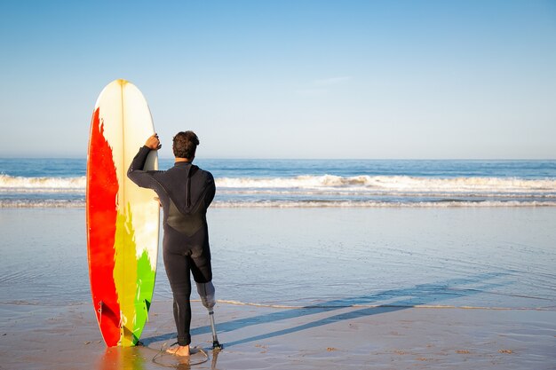 Back view of brunette surfer standing with surfboard on beach