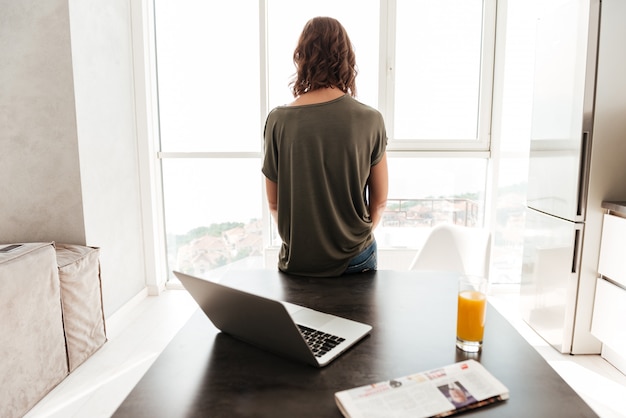 Back view of casual woman standing near table an looking at window