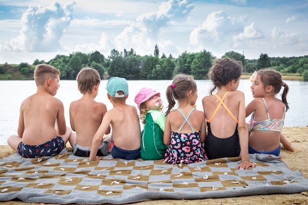 Back view children sit near the river and relax after swimming, sunbathe, eat ice cream.