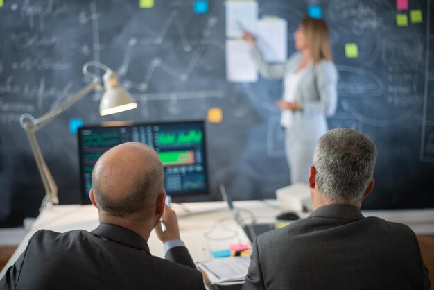 Back view of company experts working in office. Two serious man in formalwear sitting looking at woman presenting business plan and market data on board. Financial analytics, business growth concept