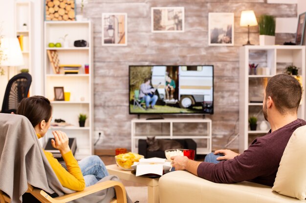 Back view of couple in living room watching a movie on the TV while eating takeaway food