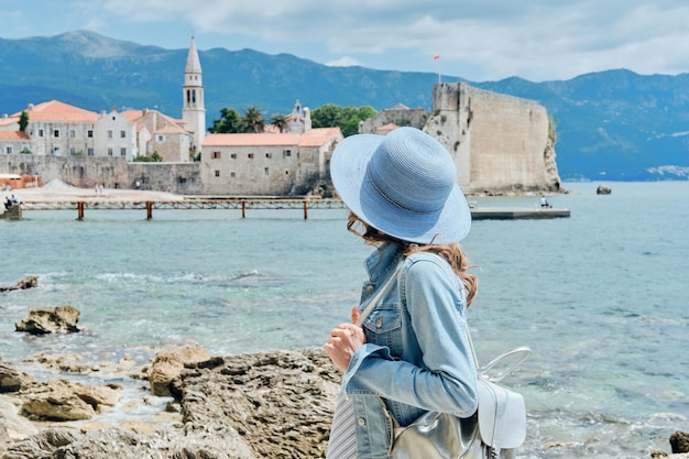 Free photo back view of curly hair woman in hat standing in blue dress looking out towards blue sea and sky in montenegro