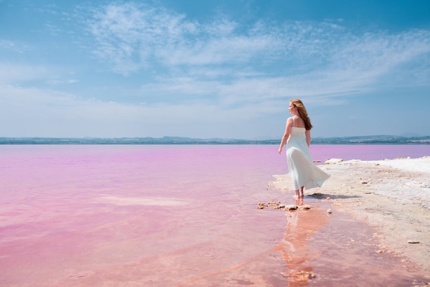 素晴らしいピンクの湖の上を歩いて白いドレスを着ているかわいい10代女性の背面図