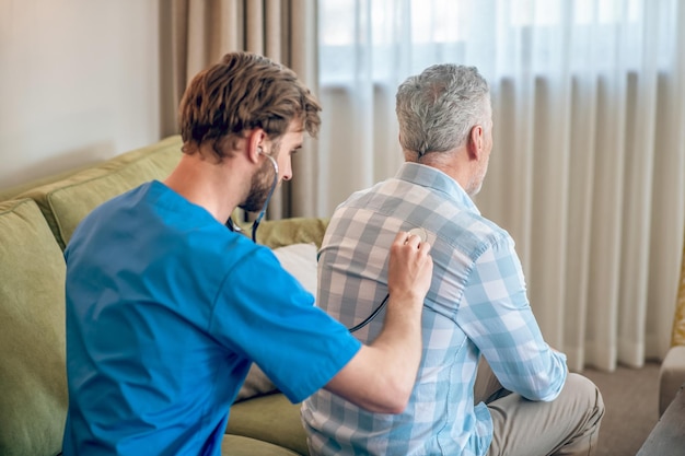 Vista posteriore di un geriatra barbuto dai capelli scuri che ausculta i polmoni di un paziente caucasico dai capelli grigi