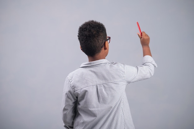 Вид сзади на темноволосого мальчика в полосатой рубашке и очках, рисующего карандашом