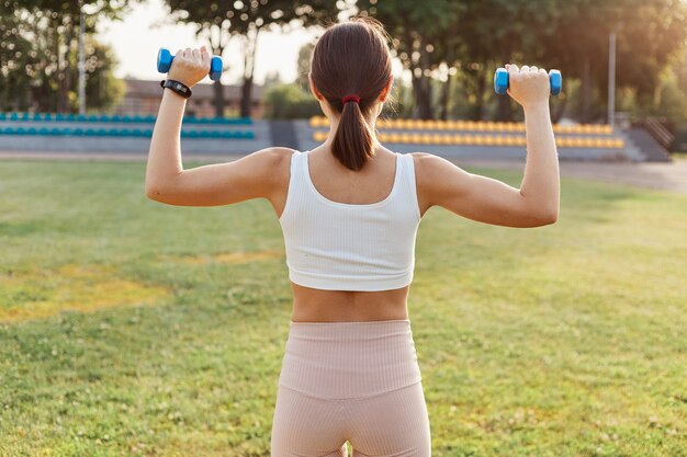 Back view of dark haired female with sporty body holding dumbbells and doing exercises on stadium, training biceps and triceps, outdoor activity, healthy lifestyle.