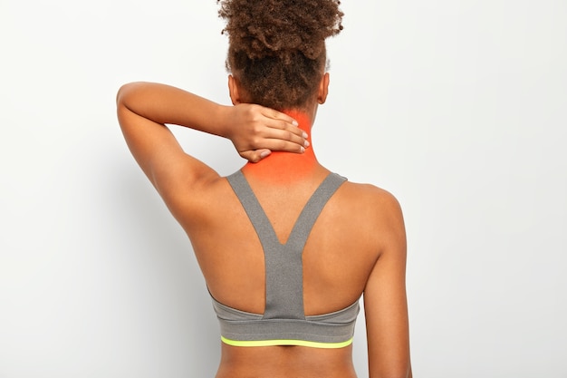 黒い肌の巻き毛の女性の背面図は首に触れ、痛みを感じ、マッサージが必要で、筋肉の損傷に苦しんでいる、灰色のトップを着て、白い壁に隔離されています