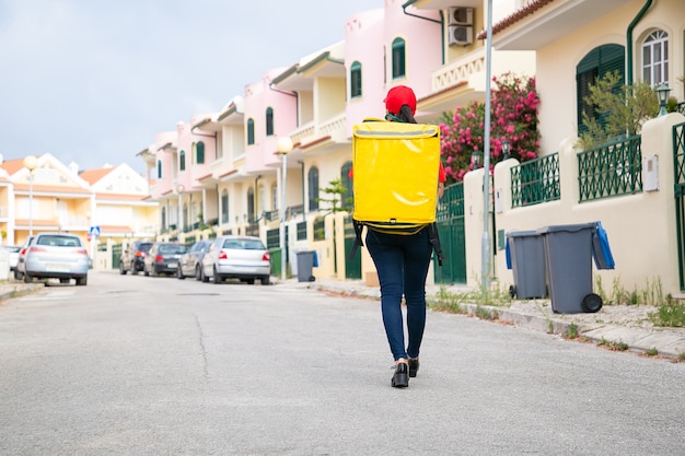 Back view of deliverywoman carrying yellow thermal bag. Female courier in red cap walking along street and delivering order.