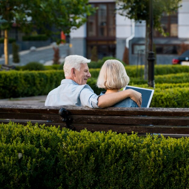 Back view of embraced older couple outdoors on bench with tablet