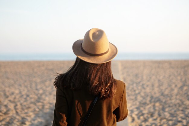 Back view of European woman in hat and coat going towards sea on boardwalk in fresh spring evening, feeling lonely