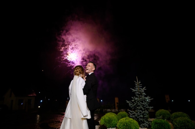 Back view of faceless bride dressed in white gown and jacket standing and watching firecrackers show on sky while her husband feeling happy and smiling on street during wedding party