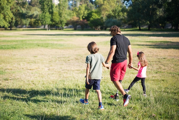 子供と一緒に歩いている障害を持つ父親の背面図。男の子と女の子と一緒に公園を散歩している機械的な脚を持つ男。障害、家族、愛の概念