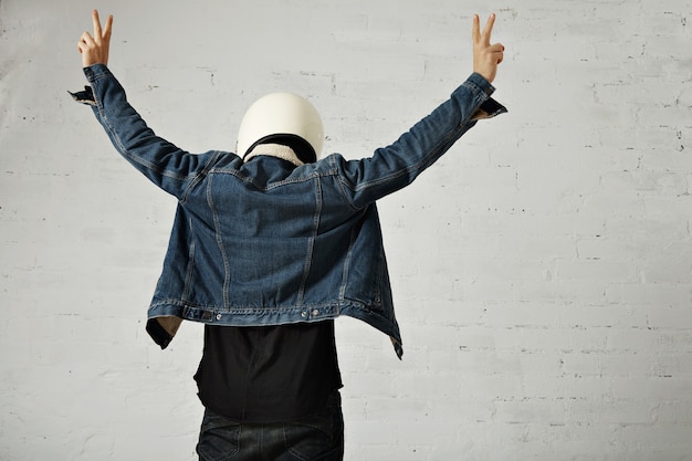 Back view on fit body of younf motocyclist wears helmet, black longsleeve henley shirt and club denim jacket with his hands up showing peace gesture